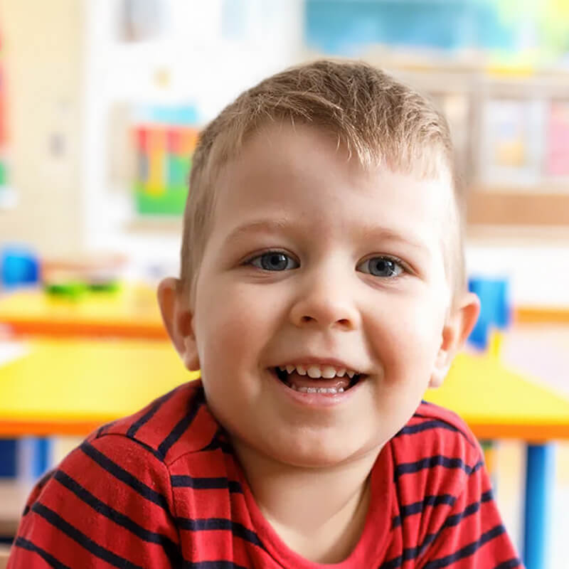 A cheerful young boy smiles at the camera, surrounded by classroom decorations and learning materials.