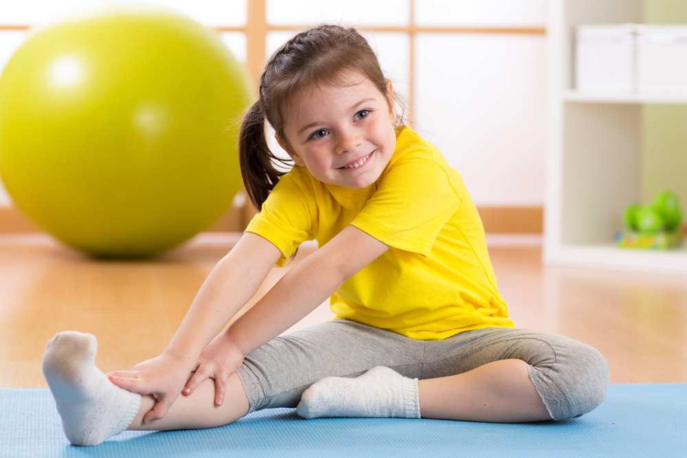 A young girl sits on a yoga mat in an indoor gym, enjoying her fitness routine in a vibrant, year-round environment.