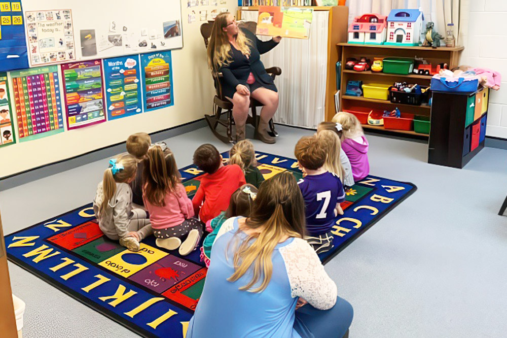 A woman reads to children in a classroom, fostering literacy and play-based learning to enhance their skills.