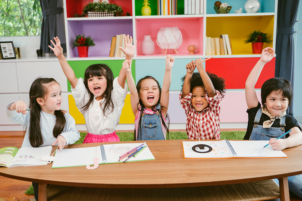 A group of children sitting at a table, hands raised in excitement, representing a welcoming and supportive care atmosphere.