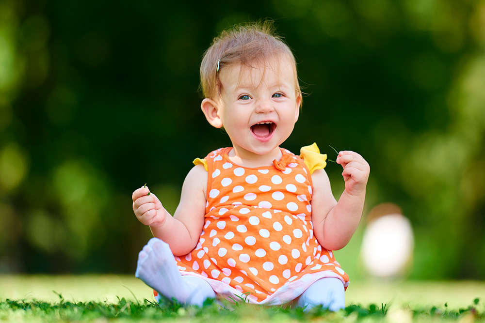 A cheerful baby girl sits on the grass with her hands up, celebrating the joy of outdoor adventures.