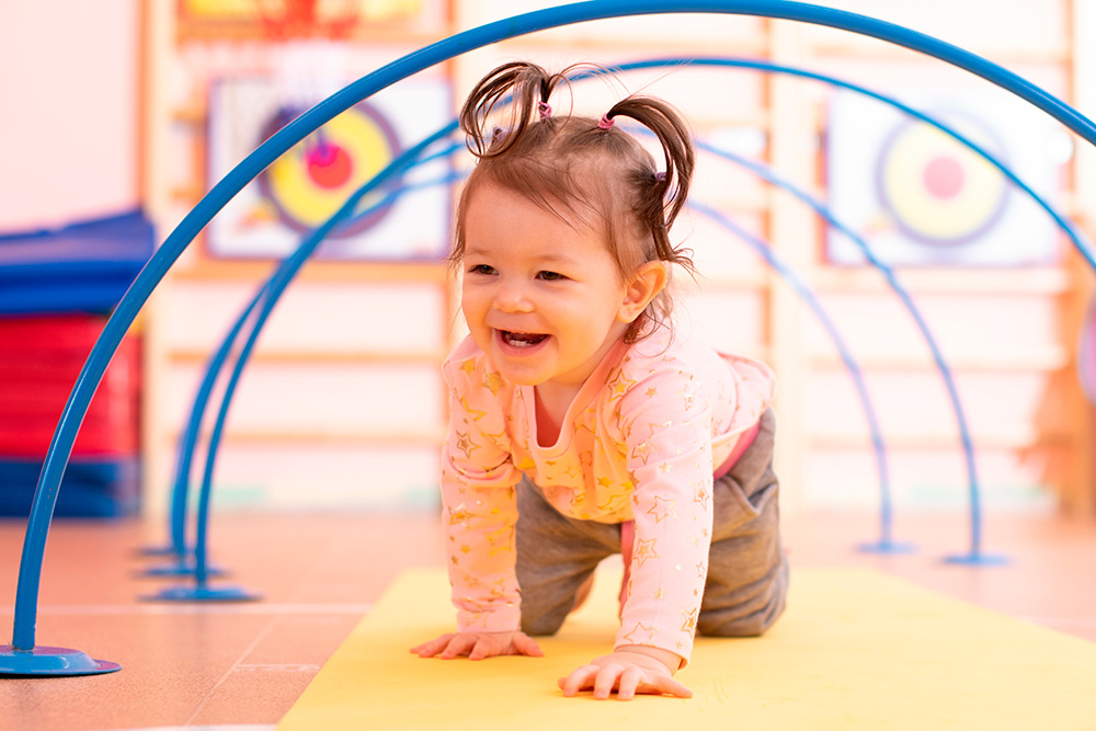 A baby girl crawls on a vibrant yellow and blue mat in an indoor gym designed for year-round activities.