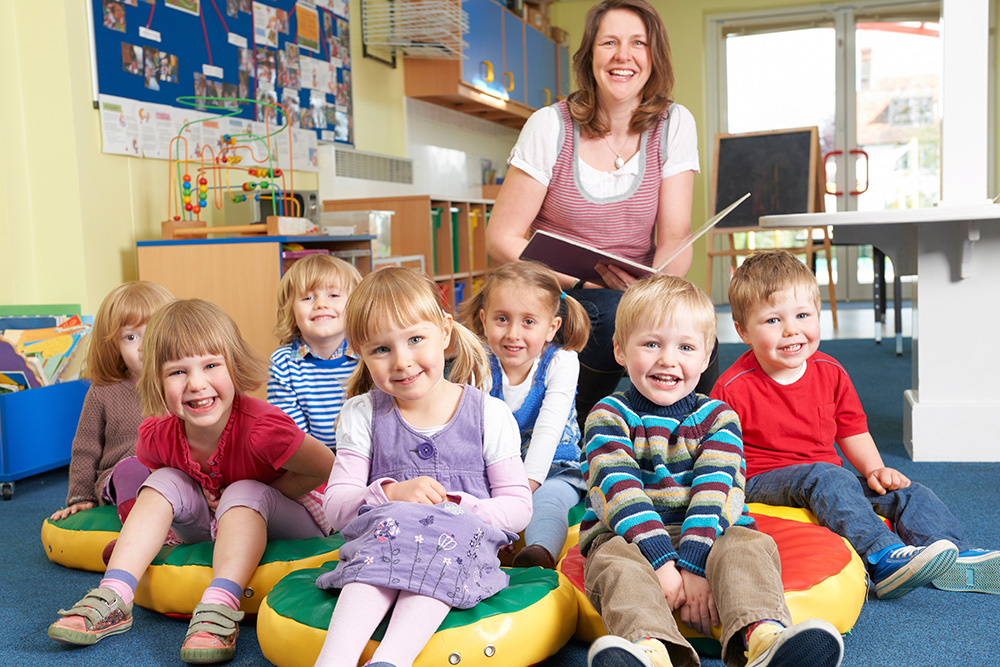 A teacher reads to a group of happy preschoolers sitting on cushions, creating a fun and engaging learning atmosphere.