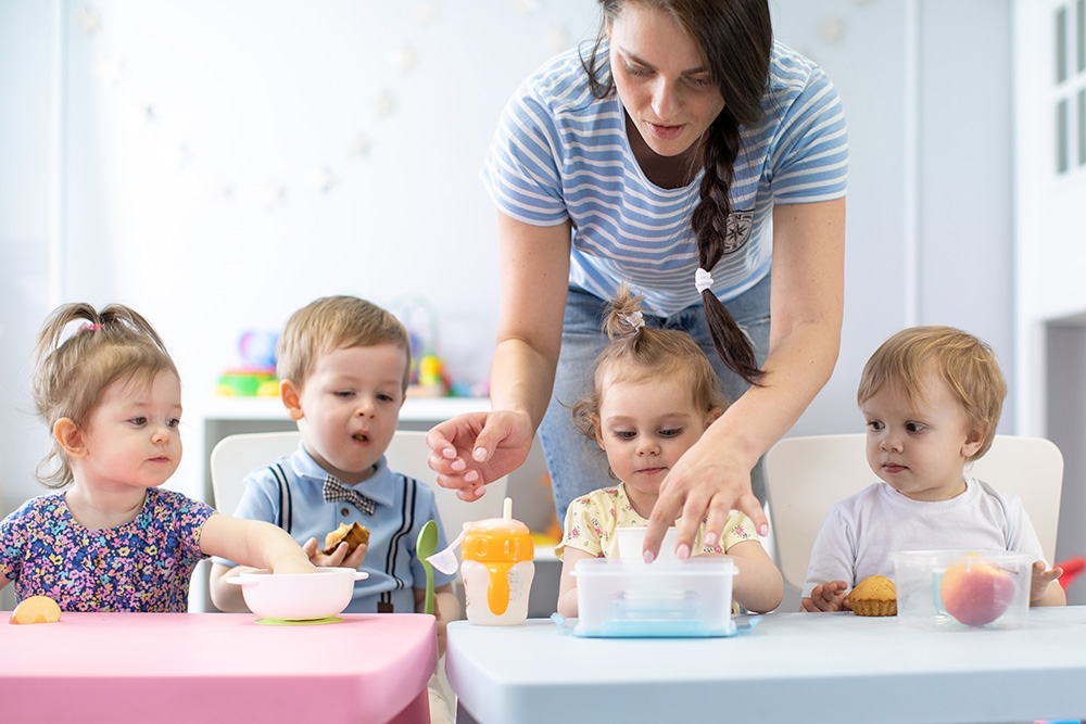 A woman serves nutritious meals to babies at a table, promoting simple and healthy mealtime practices.