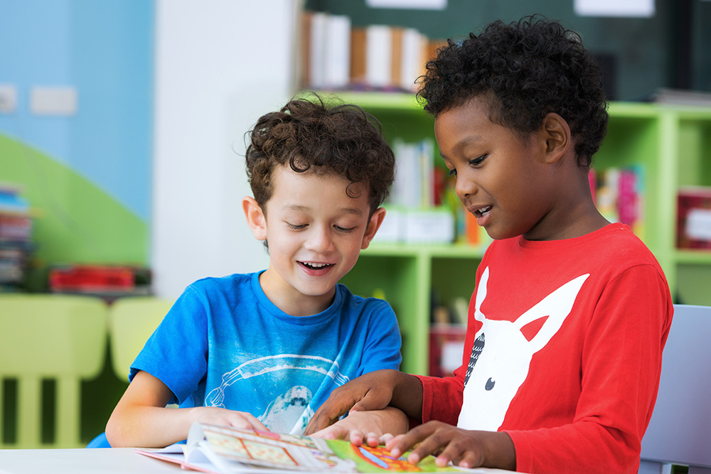 Two kids enjoy a book together in class, fostering social-emotional growth through playful interactions.