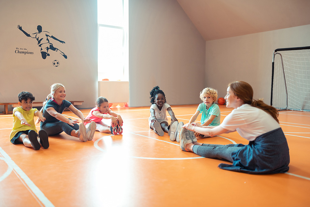 A teacher supervises children sitting on the gym floor, encouraging active participation in various activities.